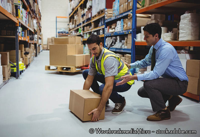 A warehouse worker  lifts a box under supervision
